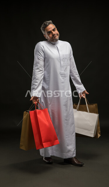 Gestures of not taking advantage of seasonal offers and discounts, the joy of buying, shopping, and acquiring supplies and gifts, a portrait of a Saudi Gulf Arab young man wearing a traditional dress, holding paper bags in his hand and looking at the camera, full body portrait, black background