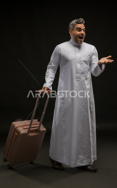 السرور والبشاشة لرؤية شخص ما, بورتريه لرجل عربي خليجي سعودي يرتدي الثوب التقليدي يمسك حقيبة السفر بإيماءات الدهشة والسعادة, الإعجاب بمكان معين, الاستمتاع بالرحلة السياحية برفقة الأصدقاء, صورة الجسم بالطول الكامل, خلفية سوداء