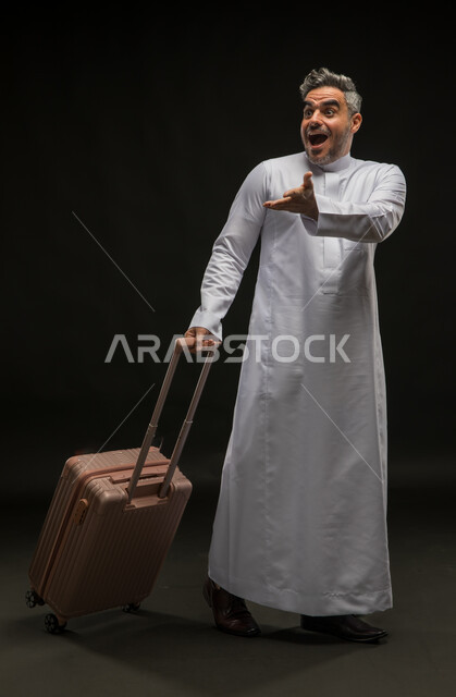 السرور والبشاشة لرؤية شخص ما, بورتريه لرجل عربي خليجي سعودي يرتدي الثوب التقليدي يمسك حقيبة السفر بإيماءات الدهشة والسعادة, الإعجاب بمكان معين, الاستمتاع بالرحلة السياحية برفقة الأصدقاء, صورة الجسم بالطول الكامل, خلفية سوداء