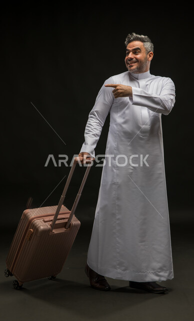 الإشارة إلى مكان معين بإيماءات الإعجاب, بورتريه لرجل عربي خليجي سعودي يرتدي الثوب التقليدي يمسك حقيبة السفر وينظر إلى شيء ما, قضاء رحلة سياحية ممتعة في وقت الإجازة الصيفية, صورة الجسم بالطول الكامل, خلفية سوداء