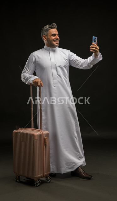القيام بالبث المباشر والتحدث مع الأصدقاء عبر وسائل التواصل الاجتماعي, مفهوم السياحة والسفر, بورتريه لرجل عربي خليجي سعودي مبتسم يرتدي الثوب التقليدي يمسك الهاتف المحمول، استخدام الأجهزة والتقنيات والتطبيقات السياحية, صورة لكامل الجسم , خلفية سوداء