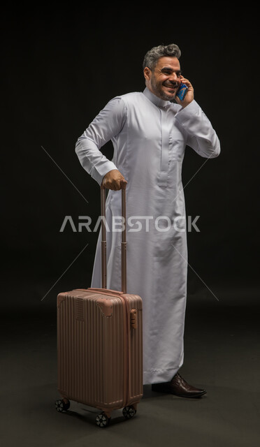 التحدث مع الأصدقاء عبر وسائل التواصل الاجتماعي, إجراء مكالمة صوتية عبر الهاتف المحمول, بورتريه لرجل عربي خليجي سعودي يرتدي الثوب التقليدي يمسك حقيبة السفر, الاستعداد لبدء الرحلة السياحية بإيماءات البشاشة والسرور, صورة الجسم بالطول الكامل, خلفية سوداء