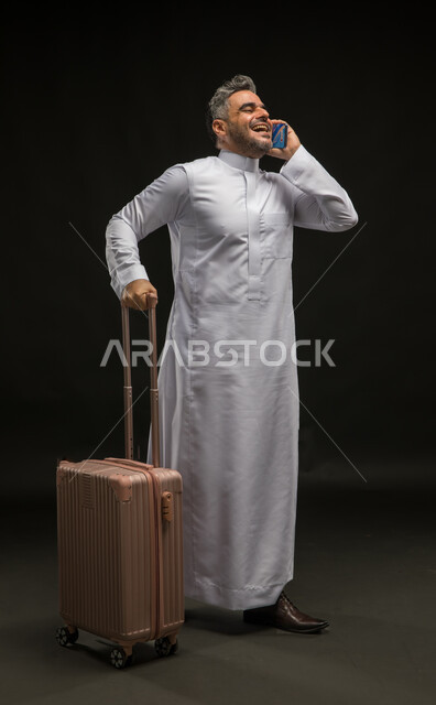 إجراء مكالمة صوتية عبر الهاتف المحمول, التحدث مع الأصدقاء عبر وسائل التواصل الاجتماعي, بورتريه لرجل عربي خليجي سعودي يرتدي الثوب التقليدي يمسك حقيبة السفر, الاستعداد لبدء الرحلة السياحية بإيماءات البشاشة والسرور, صورة الجسم بالطول الكامل, خلفية سوداء