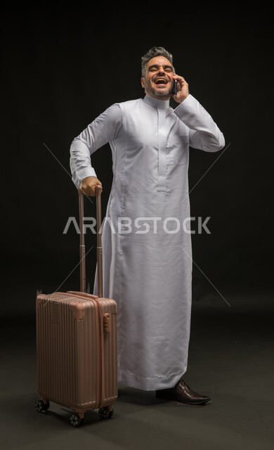 إجراء مكالمة صوتية عبر الهاتف المحمول, التحدث مع الأصدقاء عبر وسائل التواصل الاجتماعي, بورتريه لرجل عربي خليجي سعودي يرتدي الثوب التقليدي يمسك حقيبة السفر, الاستعداد لبدء الرحلة السياحية بإيماءات البشاشة والسرور, صورة الجسم بالطول الكامل, خلفية سوداء