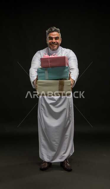 التعبير عن المحبة والألفة بين الأصدقاء, تقديم الهدايا والتهنئات، أعياد ومناسبات ومفاجآت سعيدة, بورتريه لرجل عربي خليجي سعودي مبتسم يرتدي الثوب التقليدي يحمل بيديه علب هدايا مغلفة، صورة الجسم بالطول الكامل، خلفية سوداء