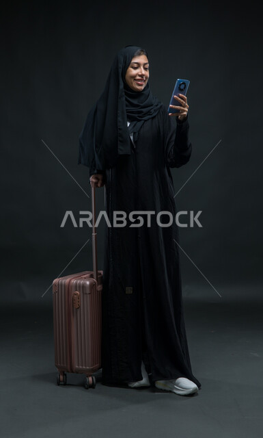 الاستفسار عن شيء ما عبر تطبيقات السياحة والسفر, بورتريه لإمرأة عربية خليجية سعودية ترتدي العباءة السوداء تمسك حقيبة السفر والهاتف المحمول بإيماءات السرور، استخدام الأجهزة والتقنيات والبرامج السياحية, صورة الجسم بالطول الكامل, خلفية سوداء