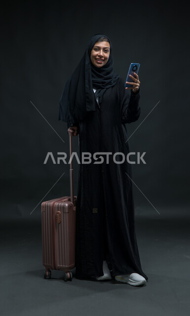 استخدام الأجهزة والتقنيات والتطبيقات السياحية, الشعور بالسرور في الخدمات المتطورة  لبرامج السياحة والسفر, بورتريه لإمرأة عربية خليجية سعودية ترتدي العباءة السوداء تمسك حقيبة السفر والهاتف المحمول بإيماءات الفرح، صورة الجسم بالطول الكامل, خلفية سوداء