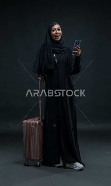الشعور بالسرور في الخدمات المتطورة  لبرامج السياحة والسفر, بورتريه لإمرأة عربية خليجية سعودية ترتدي العباءة السوداء تمسك حقيبة السفر والهاتف المحمول بإيماءات الفرح، استخدام الأجهزة والتقنيات والتطبيقات السياحية, صورة الجسم بالطول الكامل, خلفية سوداء