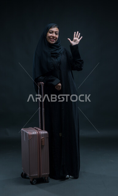 إيماءات الشكر وعدم الحاجة للمساعدة، التلويح باليد والتعبير عن الوداع,  بورتريه لإمرأة عربية خليجية سعودية مبتسمة ترتدي العباءة السوداء تمسك بيدها حقيبة السفر بتعابير الانطلاق لبدء الرحلة, صورة الجسم بالطول الكامل, خلفية سوداء