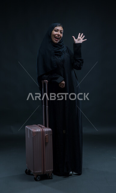 النظر إلى الكاميرا بإيماءات التعجب، التلويح باليد والتعبير عن الوداع, تعابير الانطلاق لبدء الرحلة,  بورتريه لإمرأة عربية خليجية سعودية مبتسمة ترتدي العباءة السوداء تمسك بيدها حقيبة السفر بإيماءات الشكر وعدم الحاجة للمساعدة, صورة الجسم بالطول الكامل, خلفية سوداء