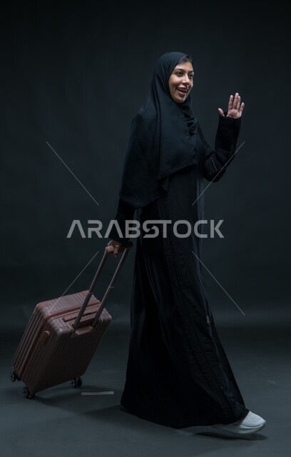تعابير الانطلاق لبدء الرحلة, التلويح باليد والتعبير عن الوداع, بورتريه لإمرأة عربية خليجية سعودية مبتسمة ترتدي العباءة السوداء تمسك بيدها حقيبة السفر بإيماءات الشكر وعدم الحاجة للمساعدة, صورة الجسم بالطول الكامل, خلفية سوداء