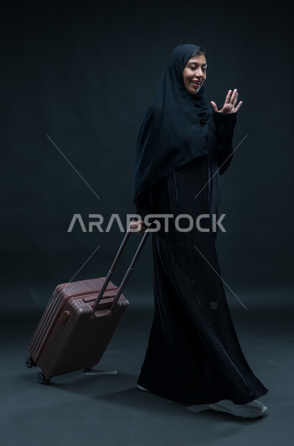 التلويح باليد والتعبير عن الوداع, تعابير الانطلاق لبدء الرحلة,  بورتريه لإمرأة عربية خليجية سعودية مبتسمة ترتدي العباءة السوداء تمسك بيدها حقيبة السفر بإيماءات الشكر وعدم الحاجة للمساعدة, صورة الجسم بالطول الكامل, خلفية سوداء