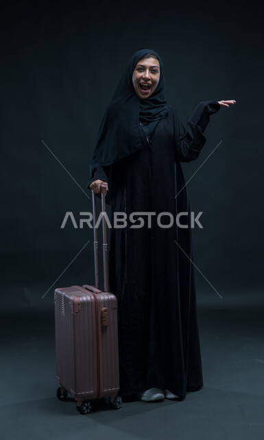 الشعور بالسعادة والفرح، السياحة في إجازة الصيف, الاستعداد لقضاء رحلة صيفية ممتعة, بورتريه لشابة عربية خليجية سعودية محجبة ترتدي العباءة السوداء تمسك حقيبة السفر تنظر إلى الكاميرا بإيماءات الحماس, صورة الجسم بالطول الكامل, خلفية سوداء.