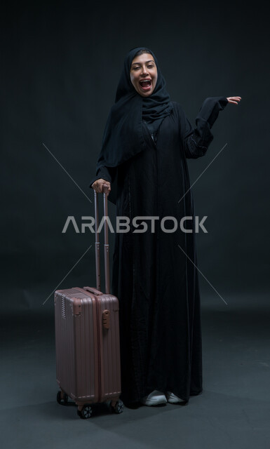 السياحة في إجازة الصيف, الاستعداد لقضاء رحلة صيفية ممتعة, بورتريه لشابة عربية خليجية سعودية محجبة ترتدي العباءة السوداء تمسك حقيبة السفر تنظر إلى الكاميرا بإيماءات الحماس, الشعور بالسعادة والفرح، صورة الجسم بالطول الكامل, خلفية سوداء.