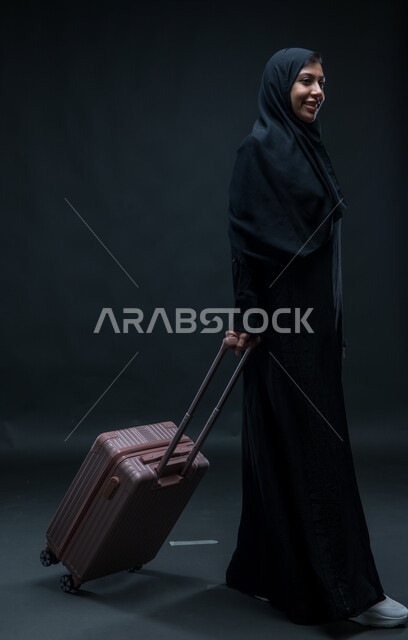 الاستعداد لقضاء رحلة صيفية ممتعة, السياحة في إجازة الصيف, بورتريه لشابة عربية خليجية سعودية مبتسمة محجبة ترتدي العباءة السوداء تمسك حقيبة السفر تسير بإيماءات السرور, الشعور بالسعادة والفرح، صورة الجسم بالطول الكامل, خلفية سوداء.