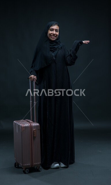 الانطلاق لقضاء رحلة صيفية ممتعة, الشعور بالسعادة والفرح، السياحة في إجازة الصيف, بورتريه لشابة عربية خليجية سعودية محجبة ترتدي العباءة السوداء تمسك حقيبة السفر تنظر إلى الكاميرا بإيماءات الحماس, صورة الجسم بالطول الكامل, خلفية سوداء.