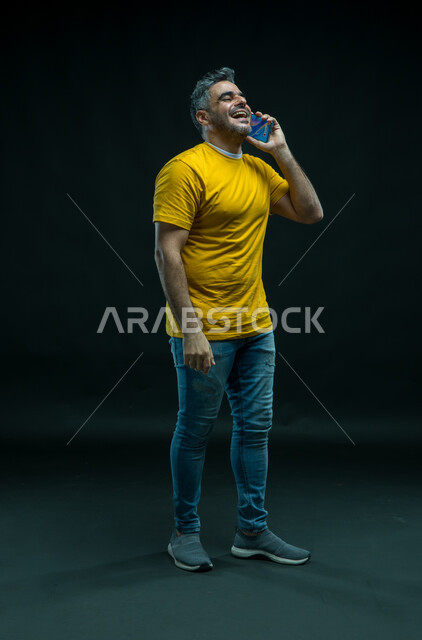 Gestures of happiness and joy, making voice calls via mobile phone, a portrait of a happy Saudi Arabian Gulf man wearing a casual outfit, standing straight and exchanging conversations with family, laughing and hanging out with friends, full body portrait, black background.
