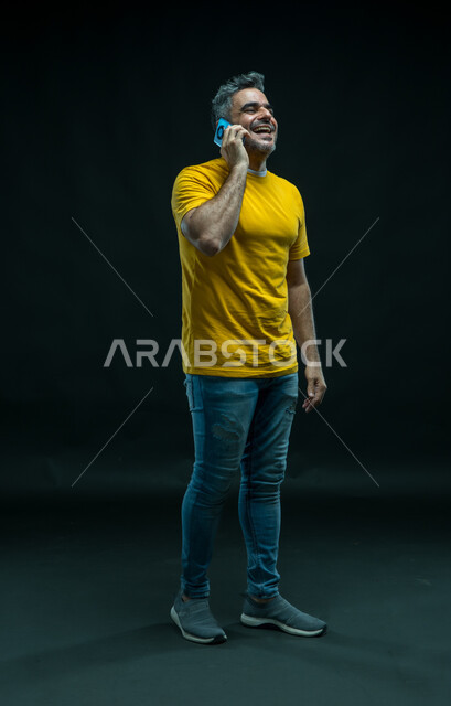 إجراء مكالمات صوتية عبر الهاتف المحمول, بورتريه لرجل عربي سعودي خليجي مسرور يرتدي الزي الكاجوال يقف باستقامة ويقوم بتبادل الاحاديث مع الاهل, الضحك والتسامر مع الأصدقاء، إيماءات السعادة والفرح, صورة الجسم بالكامل, خلفية سوداء.