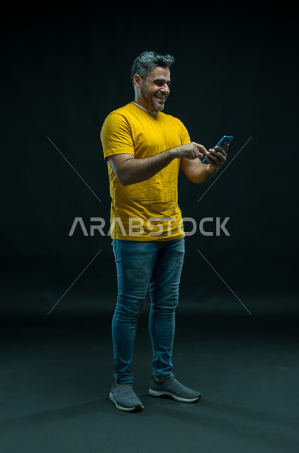 Laughing and hanging out with relatives, talking to friends via mobile phone applications, the role of modern technology in providing greater comfort for individuals, a portrait of a Saudi Gulf Arab young man wearing a casual outfit reading jokes and jokes with happy gestures, full-length body image, black background.