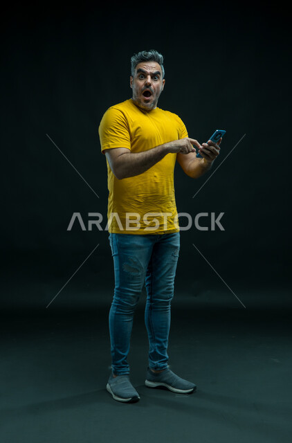 استخدام التقنيات الحديثه المتطورة، الشعور بالانزعاج لمشاهدة الأخبار المفاجئة والصادمة عبر الجوال، بورتريه لشاب عربي خليجي سعودي يرتدي الزي الكاجوال يحمل بيده الهاتف المحمول و ينظر إلى الكاميرا بتعابير الاستغراب والاندهاش، صورة الجسم بالطول الكامل، خلفيه سوداء