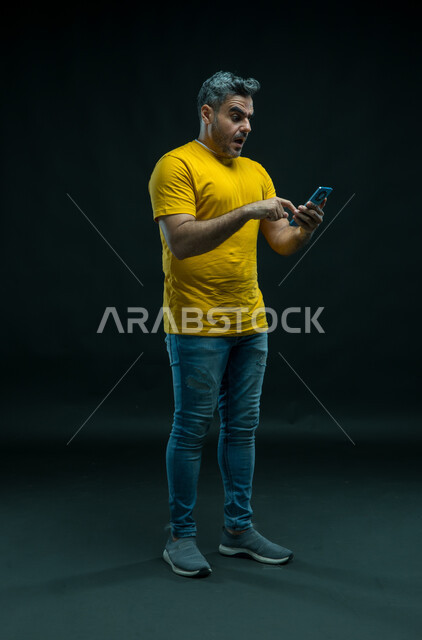 الشعور بالانزعاج لمشاهدة الأخبار السيئة الصادمة عبر الجوال، بورتريه لشاب عربي خليجي سعودي يرتدي الزي الكاجوال ينظر إلى الهاتف المحمول  بتعابير الاستغراب والاندهاش، استخدام التقنيات الحديثه المتطورة، صورة الجسم بالطول الكامل، خلفيه سوداء