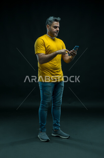 Using modern devices to develop personal skills, using a mobile phone to access information easily, completing tasks remotely, a portrait of a young Saudi Arab Gulf man wearing a casual outfit browsing social networking sites, full-length body photo, black background