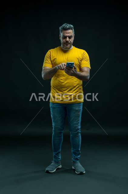 Using advanced modern technologies, chatting and friendly conversations with friends via the Internet, a portrait of a young Arab Saudi Gulf man wearing a casual outfit browsing social networking sites via a mobile phone, gestures of integration and focus, a photo of the full body, black background.