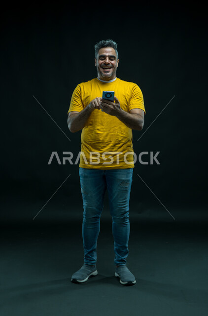 الضحك اثناء قراءة النكت والفكاهات، الدردشة والتحدث مع الاصدقاء عبر الانترنت، بورتريه لشاب عربي خليجي سعودي مبتسم يرتدي الزي الكاجوال يقوم بمتابعة اخبار وسائل التواصل الاجتماعي باستخدام الجوال، تصفح تطبيقات الهاتف المحمول، صورة للجسم بالكامل، خلفية سوداء