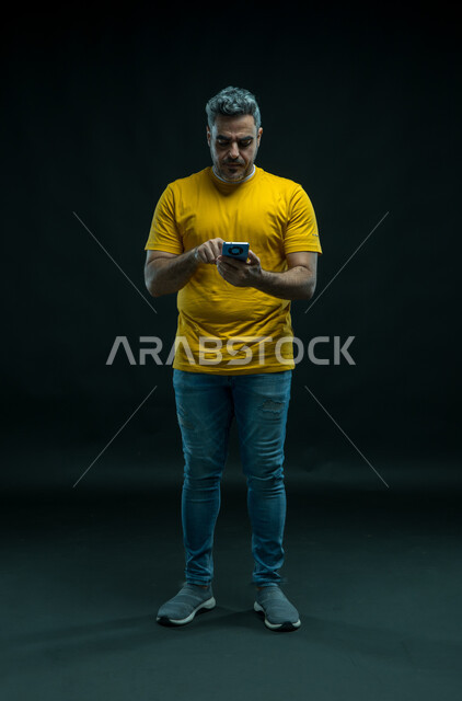 تصفح تطبيقات الهاتف المحمول، الدردشة والتحدث مع الاصدقاء عبر الانترنت، بورتريه لشاب عربي خليجي سعودي يرتدي الزي الكاجوال يقوم بمتابعة اخبار وسائل التواصل الاجتماعي باستخدام الجوال، ايماءات الاندماج والتركيز، صورة للجسم بالكامل، خلفية سوداء