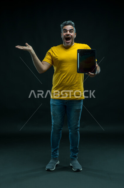 التسويق الرقمي الالكتروني، عرض شاشة اعلانية سوداء فارغة عبر التابليت، استخدام التقنيات الحديثة ،رفع اليد للاعلى بإيماءات الانفعال والاندهاش بتطبيقات الجهاز اللوحي، بورتريه لشاب عربي خليجي سعودي يرتدي الزي الكاجوال بتعابير الذهول، صورة لكامل الجسم، خلفيه سوداء