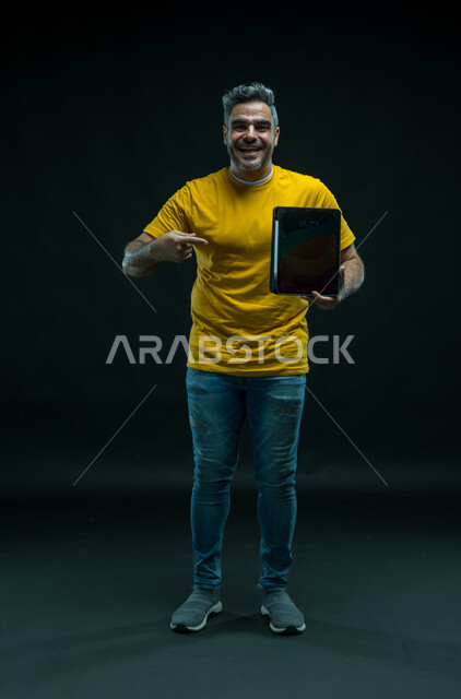 عرض شاشة اعلانية سوداء فارغة عبر التابليت، استخدام التقنيات الحديثة للتسويق الرقمي الالكتروني، الاشارة باصبع السبابه إلى الجهاز اللوحي بإيماءات السرور، بورتريه لشاب عربي خليجي سعودي يرتدي الزي الكاجوال بتعابير السعاده، صورة للجسم بالكامل، خلفيه سوداء