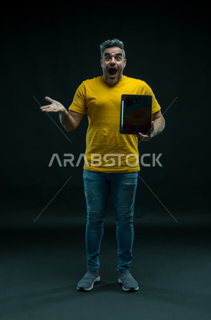 استخدام التقنيات الحديثة ،رفع اليد للاعلى بإيماءات الانفعال والاندهاش بتطبيقات الجهاز اللوحي، عرض شاشة اعلانية سوداء فارغة عبر التابليت ،التسويق الرقمي الالكتروني، بورتريه لشاب عربي خليجي سعودي يرتدي الزي الكاجوال بتعابير الذهول، صورة للجسم بالكامل، خلفيه سوداء