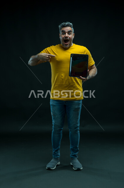 الاشارة إلى الجهاز اللوحي بإيماءات الانفعال والاندهاش، عرض شاشة اعلانية سوداء فارغة عبر التابليت، استخدام التقنيات الحديثة للتسويق الرقمي الالكتروني، بورتريه لشاب عربي خليجي سعودي يرتدي الزي الكاجوال بتعابير الذهول والسرور، صورة للجسم بالكامل، خلفيه سوداء