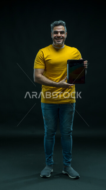 الاعجاب بتطبيقات وخدمات الجهاز اللوحي ،عرض شاشة اعلانية سوداء فارغة عبر التابليت، استخدام التقنيات الحديثة للتسويق الرقمي الالكتروني، بورتريه لشاب عربي خليجي سعودي مبتسم يرتدي الزي الكاجوال و ينظر إلى الكاميرا بإيماءات السعادة ،صورة الجسم بالكامل، خلفية سوداء