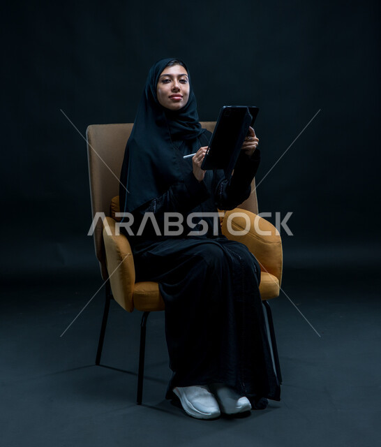 تعابير وإيماءات الثقة بالنفس, إستخدام اجهزة تقنية حديثة، تسجيل وتدوين البيانات والملاحظات، بورتريه لإمرأة عربية خليجية سعودية محجبة تجلس على كرسي مريح ترتدي العباءة السوداء تقوم بإنجاز مهام العمل عبر جهاز التابلت, خلفية سوداء