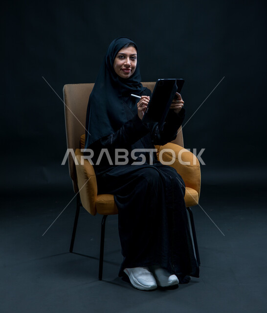 مفهوم التسوق الالكتروني الآمن، النظر إلى الكاميرا بإيماءات التركيز والسرور، تسجيل وتدوين البيانات والملاحظات، بورتريه لإمرأة عربية خليجية سعودية محجبة تجلس على كرسي مريح ترتدي العباءة السوداء تقوم بإنجاز مهام العمل عبر جهاز التابلت, خلفية سوداء