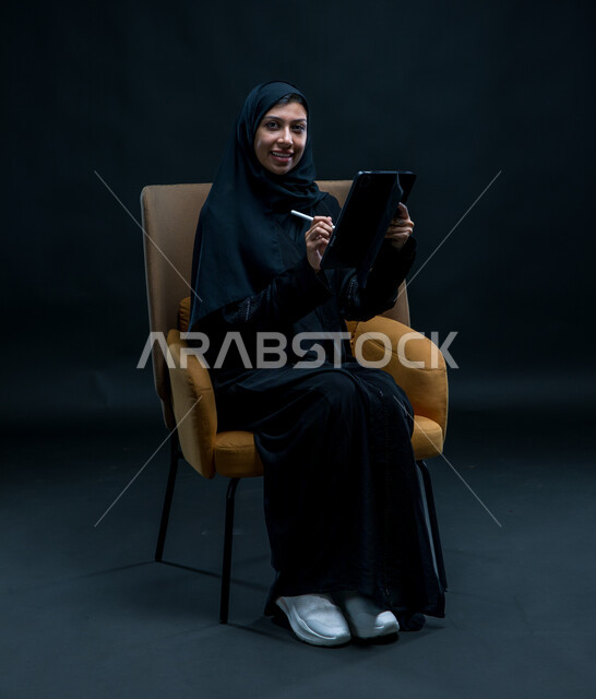 النظر إلى الكاميرا بإيماءات التركيز والسرور، مفهوم إدارة المشاريع عن بعد، تسجيل وتدوين البيانات والملاحظات، بورتريه لإمرأة عربية خليجية سعودية محجبة تجلس على كرسي مريح ترتدي العباءة السوداء تقوم بإنجاز مهام العمل عبر جهاز التابلت, خلفية سوداء