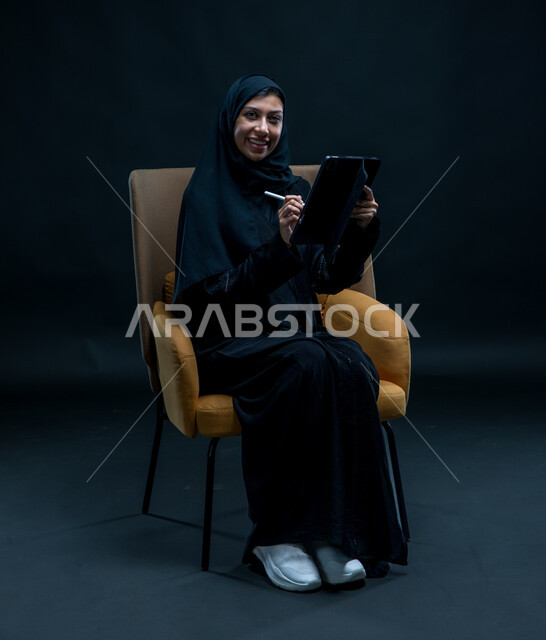 مفهوم إدارة المشاريع عن بعد، تسجيل وتدوين البيانات والملاحظات، بورتريه لإمرأة عربية خليجية سعودية محجبة تجلس على كرسي مريح ترتدي العباءة السوداء تنظر إلى الكاميرا تقوم بإنجاز مهام العمل عبر جهاز التابلت,  تعابير وإيماءات التركيز والسرور, خلفية سوداء
