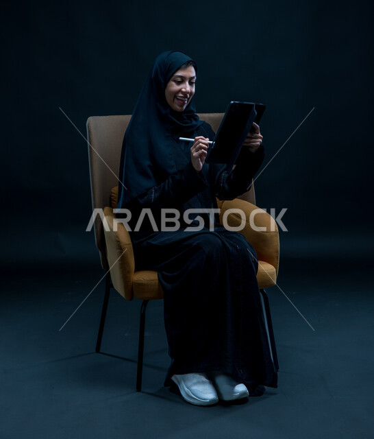 إيماءات السرور والتركيز, إستخدام اجهزة تقنية حديثة، تسجيل وتدوين البيانات والملاحظات، بورتريه لإمرأة عربية خليجية سعودية محجبة تجلس على كرسي مريح ترتدي العباءة السوداء تقوم بإنجاز مهام العمل عبر جهاز التابلت, خلفية سوداء