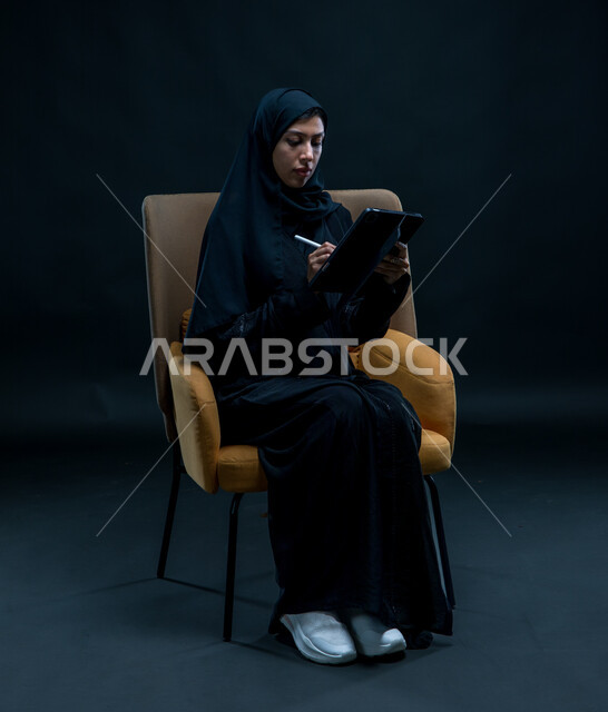 تعابير وإيماءات التركيز والاندماج, إستخدام اجهزة تقنية حديثة، تسجيل وتدوين البيانات والملاحظات، بورتريه لإمرأة عربية خليجية سعودية محجبة تجلس على كرسي مريح ترتدي العباءة السوداء تقوم بإنجاز مهام العمل عبر جهاز التابلت, خلفية سوداء
