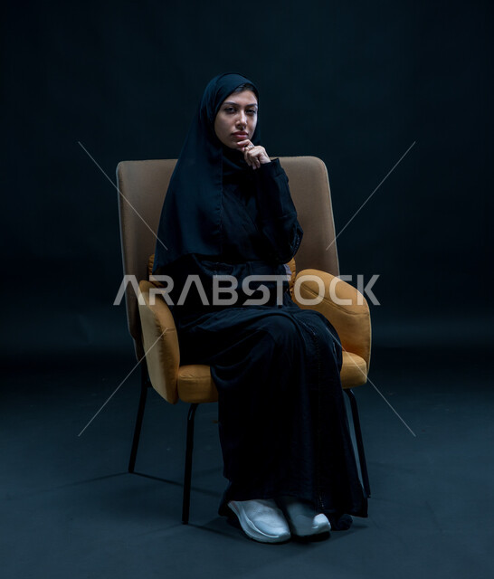 الشعور بالحيرة وعدم التأكد, إيماءات التأمل والتمعن والتركيز, بورتريه لإمرأة عربية سعودية خليجية محجبة ترتدي العباءة السوداء تجلس على كرسي مريح تضع يدها تحت رأسها بتعابير التفكير، التخطيط لحل مشكلة ما، خلفية سوداء .