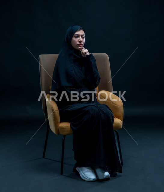 إيماءات التأمل والتمعن والتركيز, الشعور بالحيرة وعدم التأكد , بورتريه لإمرأة عربية سعودية خليجية محجبة ترتدي العباءة السوداء تجلس على كرسي مريح تضع يدها تحت رأسها بتعابير التفكير، التخطيط لحل مشكلة ما، خلفية سوداء .