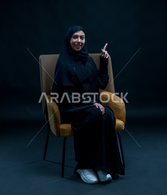 الإشاره إلى شيء ما، العناية بالجمال والشياكة، الاهتمام بالأناقة والمظهر الخارجي، بورتريه لشابة عربية خليجية سعودية محجبة تجلس على كرسي مريح ترتدي العباءة السوداء تنظر إلى الكاميرا بإيماءات الثقة بالنفس, مفهوم النعومة والأنوثة، خلفية سوداء