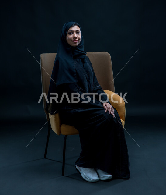 العناية بالجمال والشياكة، الاهتمام بالأناقة والمظهر الخارجي، بورتريه لشابة عربية خليجية سعودية محجبة مبتسمة تجلس على كرسي مريح ترتدي العباءة السوداء تنظر إلى الكاميرا بإيماءات الثقة بالنفس, مفهوم النعومة والأنوثة، خلفية سوداء