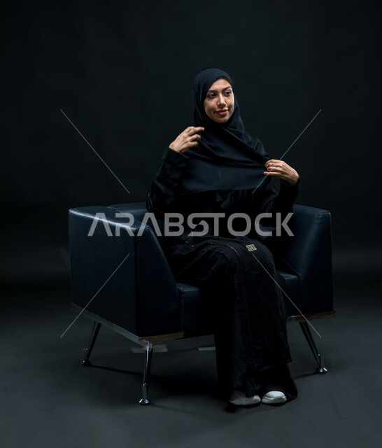 الاهتمام بالأناقة والمظهر الخارجي، تعابير وإيماءات الثقة بالنفس, بورتريه لشابة عربية خليجية سعودية محجبة تجلس على كرسي مريح ترتدي العباءة السوداء تقوم بتعديل اللباس, مفهوم النعومة والأنوثة، العناية بالجمال والشياكة، خلفية سوداء