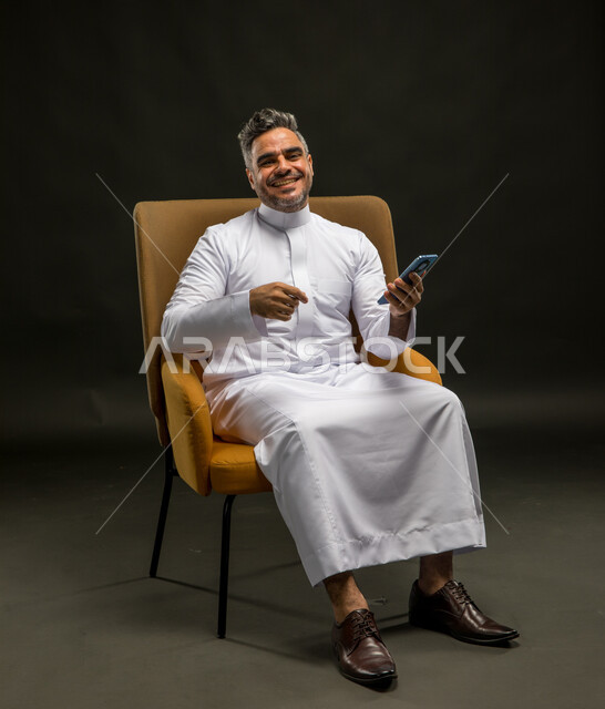 مفهوم الراحة والجلوس، بورتريه لرجل عربي خليجي سعودي يرتدي الثوب التقليدي يجلس على كرسي يحمل بيده الهاتف المحمول، قراءة الدعابات والفكاهات المضحكة، ايماءات الضحك والسرور، تصفح وسائل التواصل الاجتماعي، خلفية سوداء