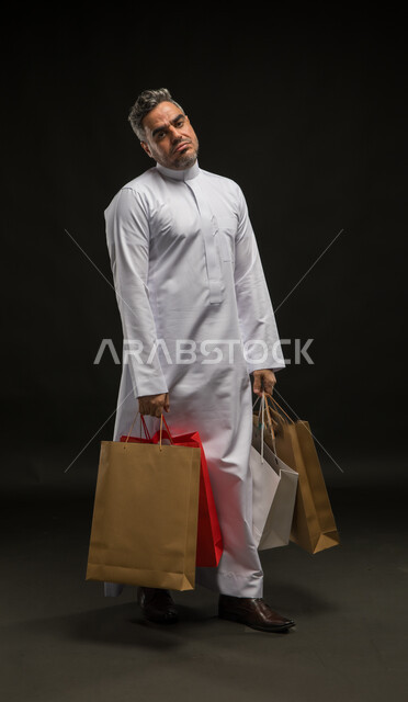 تعابير عدم الاستفادة من العروض والتخفيضات الموسمية، مفهوم الشراء والتسوق واقتناء المستلزمات، بورتريه لرجل عربي خليجي سعودي يرتدي الثوب التقليدي يحمل بيده أكياس ورقية ملونة ينظر إلى الكاميرا بإيماءت التعب, صورة الجسم بالكامل، خلفية سوداء