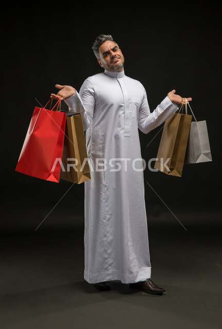 إيماءات عدم الاستفادة من العروض والتخفيضات الموسمية، مفهوم الشراء والتسوق واقتناء المستلزمات، بورتريه لرجل عربي خليجي سعودي يرتدي الثوب التقليدي يحمل بيده أكياس ورقية ملونة ينظر إلى الكاميرا بتعابير التعب والكسل, صورة الجسم بالكامل، خلفية سوداء