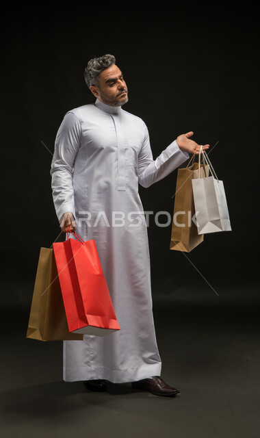 تعابير الاحساس بالتعب والارهاق من التسوق، مفهوم الشراء واقتناء المستلزمات والهدايا في التخفيضات الموسمية والجمعة البيضاء، بورتريه لرجل عربي خليجي سعودي يرتدي الثوب التقليدي يحمل بيده أكياس ورقية ملونة، صورة الجسم بالطول الكامل، خلفية سوداء