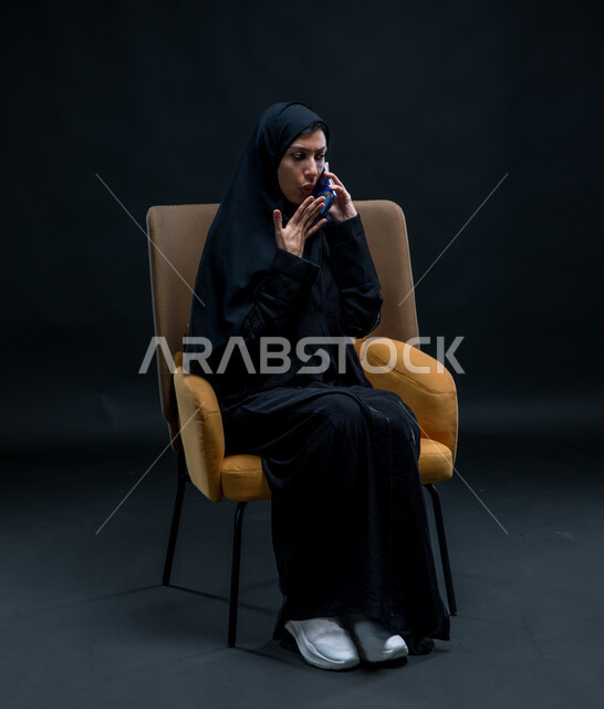 استخدام الأجهزة والوسائل التقنية الحديثة، قضاء وقت الفراغ بالتواصل مع الصديقات، إرسال الرسائل النصية والصوتية عبر الجوال، بورتريه لامرأة عربية خليجية سعودية محجبة ترتدي عباءة سوداء تجلس على كرسي مريح تقوم بإجراء مكالمة هاتفية عبر الهاتف المحمول بإيماءات الاندهاش والذهول، خلفية سوداء